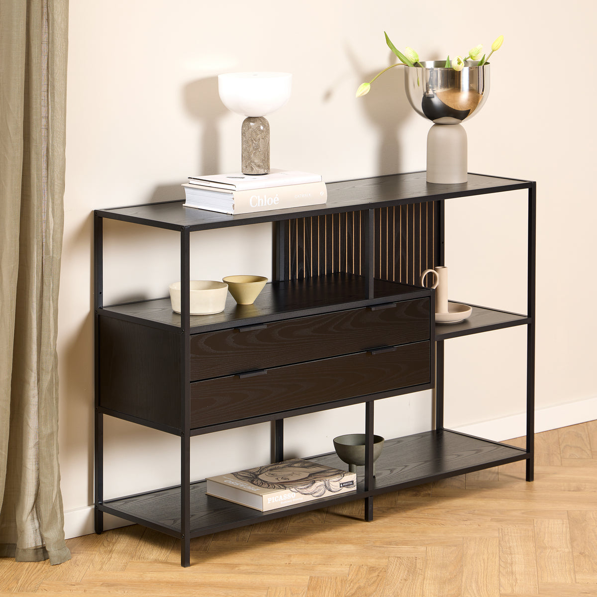 Een laag Skeleto-rek van 114 x 78, zwart melamine, tegen een lichte muur, met een lampje en een vaas met tulpen op het blad