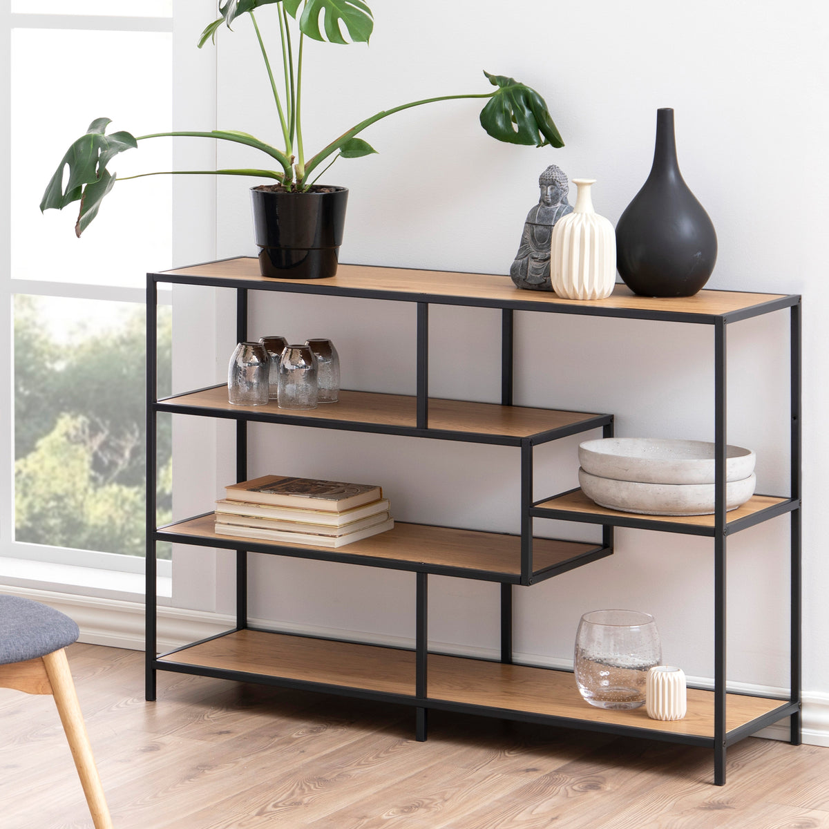 De Skeleto-boekenkast 114x78 in de kleur ‘Wild Eiken’ in een lichte woonkamer bij het raam, met een plant, vazen en boeken – een boekenkast voor in de woonkamer