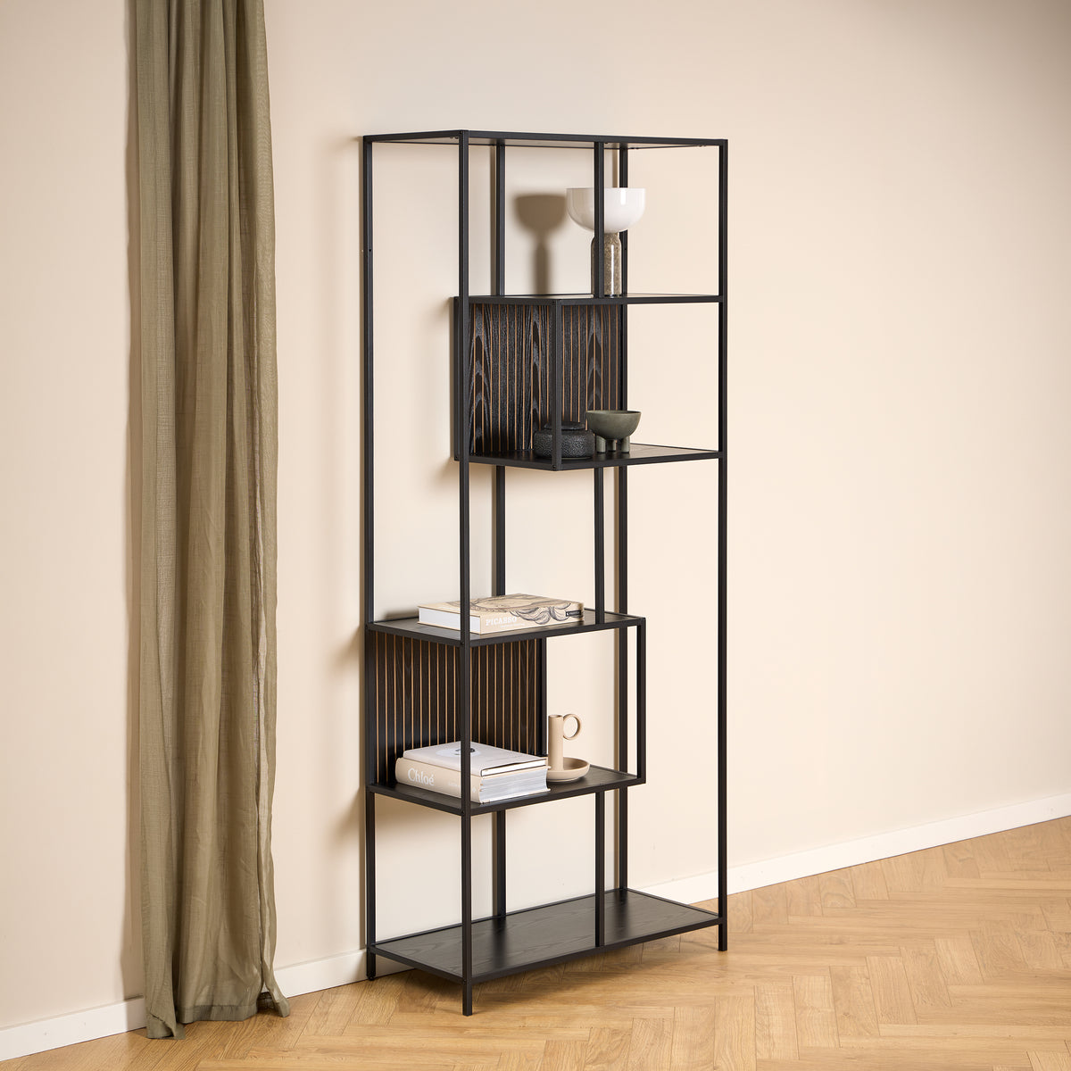 Skeleto-boekenkast 77x185, zwart melamine, tegen een lichte muur naast een linnen gordijn, met keramiek en boeken op de planken
