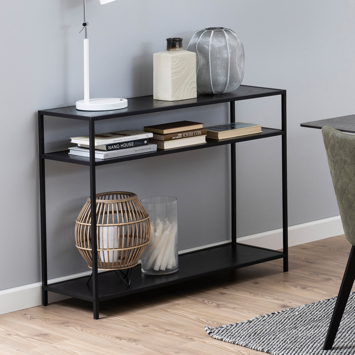 Skeleto-console 100x79, zwart melamine, met boeken, een lamp en een lantaarn op de planken, naast een stoel bij de tafel